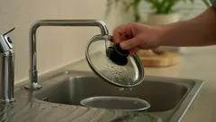 Hand hält einen Topfdeckel über Spüle, Wasser tropft ab, moderner Küchenhahn
