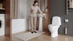 Frau zeigt auf die Toilette im modernen Badezimmer mit Waschmaschine und Waschbecken.